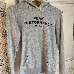 Grå hoodie från Peak Performance - Snygg ljusgrå hoodie från Peak Performance med svart tryck framtill. Klassisk ficka, huva och ribbade muddar. Perfekt för chill dagar eller när du vill ha en enkel och stilren look.