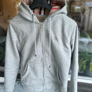 Grå hoodie från Burberry med dragkedja - Snygg grå hoodie från Burberry med klassiskt rutigt foder i huvan, dragkedja framtill och snörning. Tröjan har kängurufickor och diskret broderad logga på bröstet. Perfekt för dig som gillar stilren design med lyxig känsla.