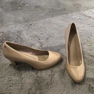 Beige pumps med klack - Stilrena pumps i blank beige färg med rund tå och låg klack. Skorna har en klassisk form och är tillverkade i syntetmaterial med en glansig finish. Perfekta för dig som gillar en clean och elegant look.