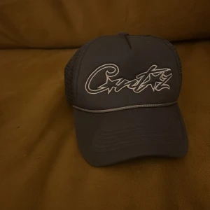 Svart meshkeps Corteiz broderad - Svart keps från Corteiz med broderad logga framtill i vitt. Klassisk trucker-modell med mesh på baksidan för extra ventilation och böjd skärm. Snygg detalj med kontrastlinje ovanför skärmen. Perfekt för en streetwear-look.