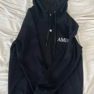 Svart AMIRI hoodie med växtmotiv - Svart hoodie från AMIRI med vit logga på bröstet och stor botanisk illustration av cannabisväxt på ryggen. Tröjan har huva med snörning, känguruficka och långa ärmar. Materialet känns mjukt och skönt, perfekt för dig som gillar streetwear med unik design.