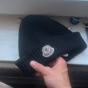 Äkta moncler mössa  - Svart ribbstickad mössa från Moncler med klassisk uppvikt kant och broderad logotyp framtill. Tillverkad i mjuk ull som håller dig varm och snygg under kalla dagar. Perfekt accessoar för dig som vill ha en stilren och exklusiv look.