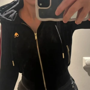 Svart velour jacka/hoodie från Moose Knuckles   - Svart velourjacka/ hoodie från Moose Knuckles med guldiga dragkedjor, huva och röda ränder längs ärmarna. Jackan har snygga detaljer som logga på bröstet och flera fickor med dragkedja. 