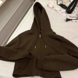 Brun hoodie med dragkedja från Zara - Säljer en brun hoodie från Zara i storlek M. Tröjan har dragkedja framtill, huva med snörning och två fickor. Modellen är croppad och har långa ärmar med ribbade muddar. Perfekt för en chill och avslappnad stil.
