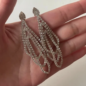 Glittriga långa örhängen - Säljer ett par statement örhängen i silverfärg med glittrande stenar. De är långa och har en öppen, geometrisk design som ger en riktigt lyxig känsla. Perfekta för att sticka ut och dra blickarna till sig. Superfina till en snygg outfit när du vill glänsa extra.