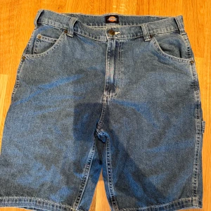 Blå jeansshorts från Dickies - Säljer ett par klassiska blå jeansshorts från Dickies med loose fit. Shortsen har flera fickor både fram och bak, bälteshällor och en cool utility-look. Perfekta för dig som gillar streetwear och avslappnad stil.