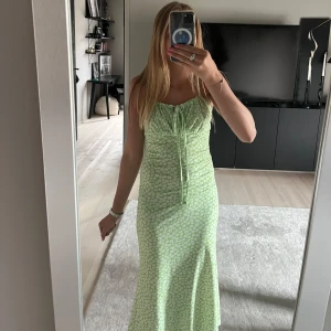 Grön blommig långklänning  - Snygg långklänning i ljusgrönt med små vita blommor. Klänningen har smala axelband, snörning framtill och en figurnära passform. Mjukt material, väldigt skön att ha på sig! Hör av dig om du har några frågor eller funderingar!🩷