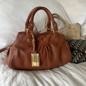 Chestnut Q Classic Baby Groove! - Så snygg klassisk chestnut färgad handväska i nyskick från Marc by Marc Jacobs i äkta läder med gulddetaljer och dubbla handtag. Väldigt snygg färg som passar till allt, den perfekta sommarväskan som rymmer mycket men är smidig och nätt. Lilla modellen Q Classic. Inga repor eller hål på väskans läder förutom ett litet märke, syns på bilderna. Otroligt välbevarad vintage modell! Perfekt för att ge en stilren touch till din outfit. Skriv privat för fler bilder. Mått 34x18x12 (undersida)