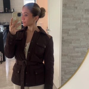 Mörkbrun quiltad jacka med bälte - Quiltad mörkbrun jacka med bälte i midjan och stora fickor framtill. Jackan har klassisk krage, knappar och markerade axelpartier. Lätt värmande jacka för vind/blås väder, passar perfekt till hösten! Storlek 34/xs