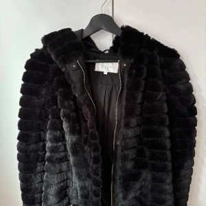 Svart pälsjacka med huva från Vila - Mysig svart pälsjacka från Vila med huva och dragkedja framtill. Jackan har ett coolt randigt mönster i pälsen och är fodrad på insidan. 