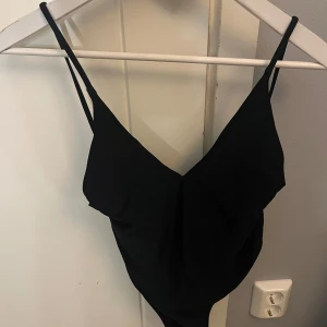 Svart baddräkt med smala axelband - Snygg svart baddräkt med smala axelband och v-ringad fram. Modellen har en enkel och stilren design med knytning i ryggen för extra passform. Perfekt för stranden eller poolen och ger en klassisk look. Oanvänd 
