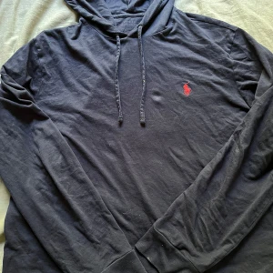 Mörkblå hoodie från Polo Ralph Lauren - Mörkblå hoodie från Polo Ralph Lauren med klassisk röd logga broderad på bröstet. Tröjan har huva med snörning och långa ärmar. Tillverkad i mjuk bomull som känns skön mot huden. Perfekt för en chill och stilren look. 