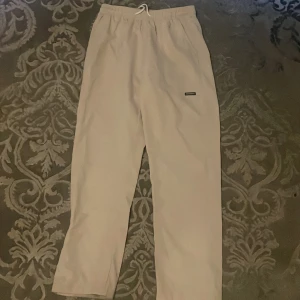 Beige polyester byxor från Progress M - Beiga polyester byxor från Progress i storlek M. Byxorna har resår och snörning i midjan för skön passform och är tillverkade i 100% polyester. Enkla och stilrena med liten logga på ena benet. Perfekta för chill dagar eller när du vill vara bekväm.