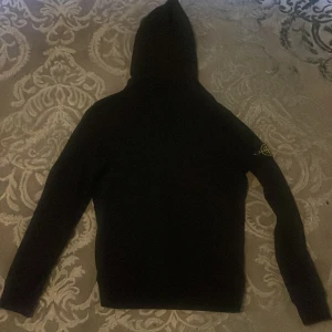Svart hoodie från Stone Island - Svart hoodie från Stone Island med klassisk patch på vänster ärm. Tröjan har huva och långa ärmar, tillverkad i mjuk bomull. Perfekt för dig som gillar streetwear och vill ha en stilren look med ikonisk logga.