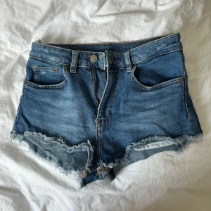 Blåa jeansshorts - Snygga blå jeansshorts med rå, fransig kant och klassisk femficksdesign. Shortsen har normal passform och är perfekta för varma dagar. De har bälteshällor, knapp och dragkedja framtill samt slitna detaljer för en avslappnad look