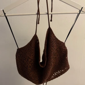 Brun virkad croppad linnetopp - Säljer en brun croppad linnetopp med virkade detaljer och tunna knytband i nacken. Toppen är ärmlös och har en snygg öppen rygg. 