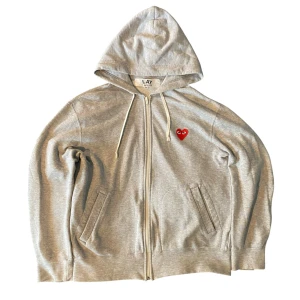 Grå zip hoodie Comme des Garçons Play - Grå zip hoodie från Comme des Garçons Play med dragkedja och huva. Ikoniskt rött hjärta med ögon broderat på bröstet. Mjuk bomullskvalitet, två fickor fram och vita snören i huvan. Storlek M men sitter mer som S, cropped passform.