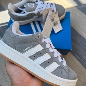 Gråa Adidas Campus sneakers - Snygga Adidas Campus sneakers i grå mocka med vita detaljer och klassiska tre ränder på sidan. Skorna har vit sula och vita skosnören, samt en gummisula undertill. Perfekta för en stilren och avslappnad look.