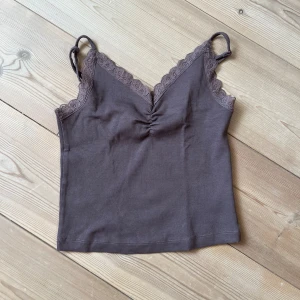 Brun ribbad linnetopp från Bik Bok - Supersöt brun ribbad linnetopp från Bik Bok med spetsdetaljer längs v-ringningen och smala axelband. Toppen har en lätt rynkning framtill och är perfekt för att skapa en chill och trendig look. Materialet är mjukt och stretchigt för extra komfort.