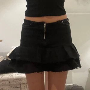 Svart volangkjol i jeans med dragkedja - Säljer en svart kort jeanskjol med volanger och råa kanter. Kjolen har framfickor, bakfickor och en synlig dragkedja framtill. 