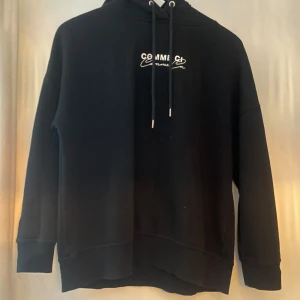 Svart hoodie från Amisu med tryck - Svart hoodie från Amisu med vit text 'Comme Ci Comme Ca' tryckt både fram och bak. Är använd och har lite slitage på trycket som kan ses på bilden. Den har både tryck på framsidan och på ryggen