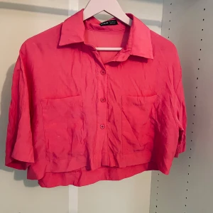 Rosa croppad skjorta med fickor - Snygg rosa croppad skjorta från SHEIN med klassisk krage, knäppning framtill och två stora bröstfickor. Skjortan har kortare passform och är tillverkad i ett lätt material som passar perfekt till varmare dagar.