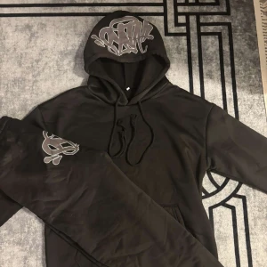 Syna World tracksuit svart  - Svart hoodie med stor ficka framtill och coolt graffiti-inspirerat tryck i silver på huvan. Tröjan har snörning i huvan och är tillverkad i mjukt material, perfekt för en streetwear-look. Långärmad och med avslappnad passform.