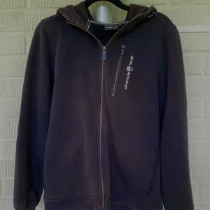 Svart hoodie från Sail Racing - Svart hoodie från Sail Racing i storlek 170 med dragkedja hela vägen, huva och en bröstficka med logga. Tröjan har långa ärmar och ribbade muddar. Perfekt för dig som gillar sportig och clean stil. Materialet känns mjukt och bekvämt, troligen bomull eller polyester.