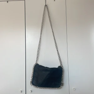 Svart Tiamo väska - Svart axelväska från Tiamo i skinnimitation med grova silverfärgade kedjor runt kanterna och som axelrem. Väskan har dragkedja upptill, extra fack med dragkedja på baksidan och magnetknappar. 