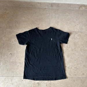 Svart t-shirt från Polo Ralph Lauren - Klassisk svart t-shirt från Polo Ralph Lauren med broderad logga på bröstet. T-shirten är i mjuk bomull och har en normal passform med korta ärmar och rund hals. Perfekt basic-plagg med stilren look. Storlek M Perfekta för de varma sommarkvällarna