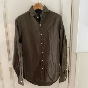 Olivgrön skjorta Polo Ralph Lauren S - Snygg olivgrön skjorta från Polo Ralph Lauren i slim fit-modell. Skjortan har klassisk krage, vita knappar och långa ärmar. Tillverkad i mjuk bomull som känns skön mot huden. Perfekt för dig som gillar stilrena och tidlösa plagg.