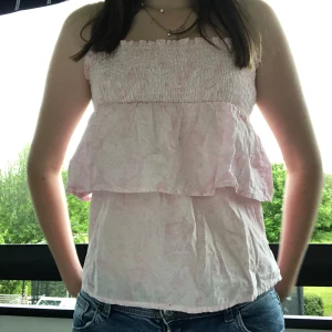 Ljusrosa volangtopp - Supersöt ljusrosa (mönstrad) linnetopp med smock upptill och volanglager längs magen. Toppen har smala och justerbara axelband, perfekt för varma dagar. Snygg att matcha med jeansshorts eller kjol. Toppen är knappt använd! 