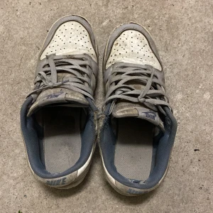 Nike gråvita sneakers med snörning - Nike sneakers i grått och vitt med klassisk snörning och perforerad tå. Skorna har lågt skaft och blågrå insida, samt tydlig Nike-logga på plösen och hälen. Ovandelen är i skinn och syntet, perfekt för dig som gillar en sportig och avslappnad stil. Går att tvätta men har inte gjort det