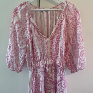 Rosa och vit kortklänning med v-ringning - Supersöt kortklänning från Zara i vitt och rosa med blommigt mönster. Klänningen har v-ringning, resår i midjan och puffiga trekvartsärmar. Perfekt för varma dagar när du vill känna dig både chill och snygg. Populär klänning!!