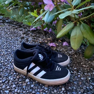 Svarta Adidas sneakers med vita ränder - Klassiska svarta Adidas sneakers med de ikoniska tre vita ränderna på sidorna och vit sula. Skorna har snörning och en låg profil, perfekt för en avslappnad streetstyle. Materialet är troligtvis mocka eller syntet, med rund tå och platt sula.