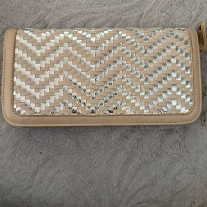 Beige och silver plånbok med mönster - Snygg plånbok i beige och silver med vävt zickzackmönster. Plånboken har dragkedja runtom och flera fack för kort och mynt. Perfekt storlek för att ha med sig överallt och ger en riktigt fräsch vibe till din look. Helt oanvänd.