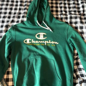 Grön Champion hoodie med logga - Säljer en grön hoodie från Champion i storlek medium. Tröjan har stor vit och gul Champion-logga framtill, huva med vita snören och en klassisk känguruficka. Perfekt för dig som gillar streetwear och vill ha en skön och snygg tröja.