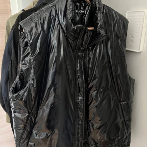 Svart glansig dunväst från Acne Studios - Säljer en svart, glansig dunväst från Acne Studios med hög krage och dragkedja framtill. På ryggen sitter en gul patch med ett ikoniskt ansikte. Västen har en oversized passform och är perfekt för lager-på-lager-stil.