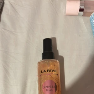 La Rive Sparkling Rose Body Mist - La Rive Sparkling Rose Body Mist med skimrande guldglitter i en genomskinlig plastflaska. Doften är rosig och fräsch, perfekt för dig som gillar blommiga toner. Flaskan har svart spraykork och rosa detaljer på etiketten.