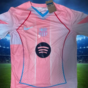 Rosa FC Barcelona Nike fotbollströja - Snygg rosa FC Barcelona fotbollströja från Nike med Spotify-logga på bröstet. Tröjan har korta ärmar, V-ringning med blå och vita detaljer samt tunna vita linjer och röda kanter på framsidan. Materialet är lätt och andas, perfekt för match eller träning.
