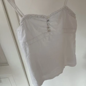 Vit linnetopp - Söt vit linnetopp med justerbara axelband och söta knappar framtill. Toppen har en fin volangkant längs urringningen och är 100% bomull. Perfekt för varma dagar och enkel att matcha med jeans eller kjol. Strl XS/S. Pyyyytteliten fläck som syns på sista bilden (typ omöjlig att märka).