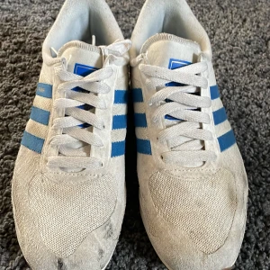 Adidas LA Trainer sneakers - Säljer ett par klassiska Adidas LA Trainer sneakers i vitt och blått. Skorna har den ikoniska tre-randiga designen och en sula med nitar för extra grepp. Perfekta för en retro look!