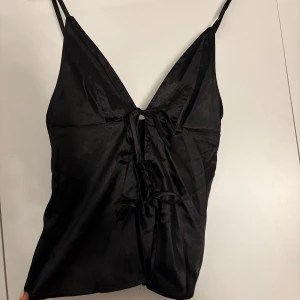 Svart satinlinne med knytning - Elegant svart linne i satin med smala axelband och dekorativ knytning framtill. Perfekt för en stilren look.