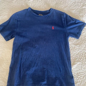 Blå t-shirt från Ralph Lauren - Säljer en klassisk blå t-shirt från Ralph Lauren med det ikoniska röda broderade logotypen på bröstet. T-shirten har en rund hals och korta ärmar, perfekt för en avslappnad stil.