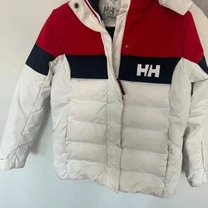 Vit och röd dunjacka från Helly Hansen - Säljer en snygg vit och röd dunjacka från Helly Hansen med marinblå detaljer. Jackan har en dragkedja framtill och är långärmad. Perfekt för kyliga dagar med sin varma och bekväma design. Helly Hansen-logga på bröstet och ryggen.