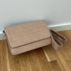 Beige axelväska med krokodilmönster - Snygg beige axelväska med krokodilmönster och justerbar axelrem. Väskan har en praktisk dragkedja inuti för säker förvaring. Perfekt för att ge din outfit en stilfull touch.