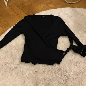 Svart omlottopp från Gina Tricot - Säljer en stilren svart omlottopp från Gina Tricot. Toppen har långa ärmar och knytning i sidan för en justerbar passform. Perfekt för både vardag och festliga tillfällen.