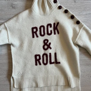 Vit polotröja från Zadig & Voltaire rock&roll - Säljer en stilren vit polotröja från Zadig & Voltaire med texten 'Rock & Roll' i vinrött på framsidan. Tröjan har en hög ribbad krage och knappar på axeln för en unik look. Perfekt för att hålla sig varm och trendig.