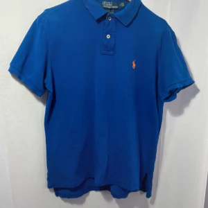 Blå pikétröja från Ralph Lauren - Säljer en klassisk blå pikétröja från Ralph Lauren med den ikoniska loggan broderad i orange på bröstet. Tröjan har korta ärmar och knappar vid kragen. Perfekt för en stilren look!