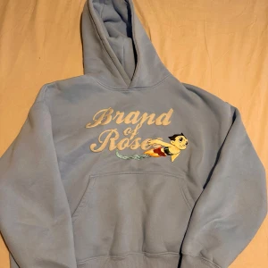 Ljusblå hoodie från Brand of Rose - Säljer en ljusblå hoodie från Brand of Rose med en cool tecknad figur på framsidan. Hoodien har en stor ficka framtill och en bekväm huva. Perfekt för en avslappnad stil! 💙